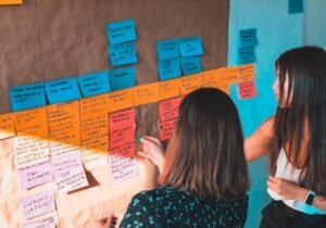Two women engaged in collaborative planning using colorful Post-it notes on a board in a modern office space.